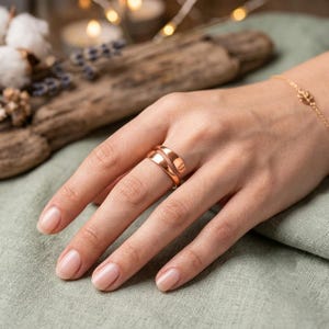 Könnte beinhalten: Ein polierter Ring aus Roségold im Bypass-Design mit zwei überlappenden Bändern, die sich um den Finger legen. Das Metall hat eine glatte, reflektierende Oberfläche und einen warmen metallischen Farbton, der eine fließende Form bildet.