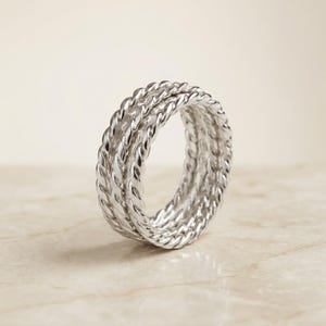 Könnte beinhalten: Ein silberner Ring mit einem dreifachen Banddesign, bei dem jedes Band eine gedrehte Seilstruktur aufweist. Die metallische Oberfläche ist poliert und reflektierend und zeigt ein durchgehendes Spiralmuster, das den gesamten Umfang umschließt.
