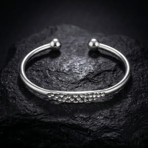 Könnte beinhalten: Ein silberfarbenes Armband in offener Form mit einem geschwungenen Mittelteil, das mit mehreren klaren, funkelnden Steinen besetzt ist. Das Armband hat eine polierte metallische Oberfläche und endet in zwei glatten, runden Kugeln.