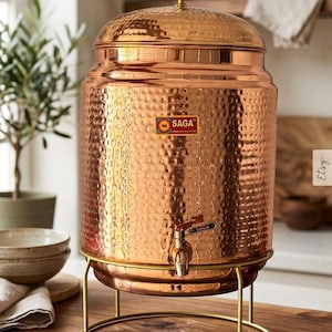 Könnte beinhalten: Ein Kupfer-Wasserspender mit gehämmertem Muster, einem Wasserhahn und einem Deckel. Der Spender steht auf einem goldfarbenen Ständer und hat das "SAGA"-Logo. Er steht auf einer Holzoberfläche.