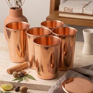 Può includere: Un set di cinque bicchieri da bere color rame con finitura lucida. I bicchieri cilindrici sono disposti su una superficie chiara, insieme a un piccolo blocco di legno, spezie e una fetta di limone. È visibile anche un coperchio color rame.
