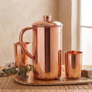 Könnte beinhalten: Ein Kupferkrug mit Deckel und Griff, zusammen mit zwei Kupferbechern. Der Krug und die Becher werden auf einem Holztablett präsentiert. Die Kupferartikel haben eine glänzende, reflektierende Oberfläche.