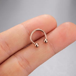 Könnte beinhalten: Ein kreisförmiges Barbell-Piercing aus roségoldfarbenem Metall mit einer glatten, polierten Oberfläche. Der hufeisenförmige Ring ist mit zwei kleinen, abschraubbaren Kugeln an den Enden versehen, die einen sicheren Verschluss des gebogenen Stabs bieten.