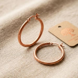 Puede incluir: Un par de pendientes de aro en oro rosa con un acabado liso y pulido y cierres de bisagra. Los pendientes son de forma circular y presentan un diseño sencillo y continuo. Una pequeña etiqueta de papel marrón con un logotipo de árbol al lado.
