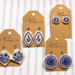 May include: A collection of blue and white earrings on display. The earrings feature various designs, including floral patterns and botanical motifs. Each pair is attached to a brown tag with the text "Lilyvale Trinkets Co."