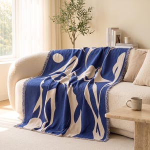 Puede incluir: Una manta tejida con un fondo azul intenso que presenta grandes formas orgánicas abstractas de color crema y una forma circular. La manta rectangular tiene bordes con flecos y está colocada sobre un sofá curvo de color claro.