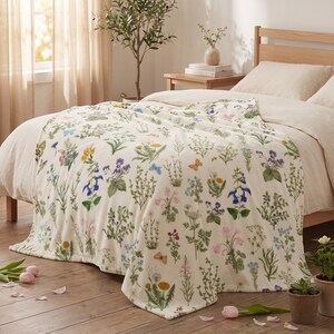 Puede incluir: Una manta de color crema con un estampado botánico repetitivo de diversas flores silvestres, follaje verde y pequeñas mariposas en tonos azules, amarillos y rosas. La tela suave está colocada sobre una cama con cabecero de madera.