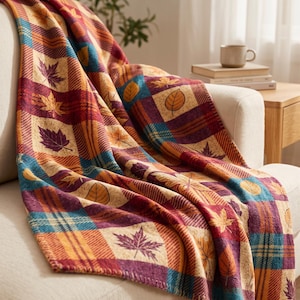 Puede incluir: Una manta tejida con un patrón de cuadros que alterna cuadrados en morado oscuro, azul verdoso y crema. El diseño está decorado con varios motivos de hojas en tonos naranja, amarillo y morado, creando una apariencia texturizada.