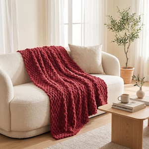 May include: A deep red chunky knit throw blanket with a textured bobble pattern draped over a cream-colored curved sofa. The blanket features a thick, soft yarn construction with a repeating raised circular design across its entire surface.