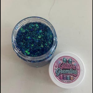 May include: A clear plastic jar filled with dark blue and teal chunky glitter. The white lid features a circular label with a pink border, a blue bow, and the text Glitter Gremlins Made to Sparkle. The text Mermaid Mischief (Blue) is below.