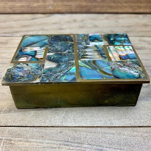 May include: A rectangular box with a brass-toned metal base and a lid inlaid with iridescent abalone shell tiles. The shell pieces feature swirling patterns of blue, green, and pearlescent white, creating a mosaic surface on the top of the container.