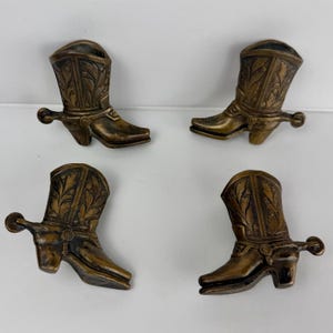 May include: A set of four bronze-toned cowboy boot figurines, each featuring intricate embossed leaf patterns on the shaft and a small spur attached to the heel. These decorative items are arranged in a two-by-two grid formation.