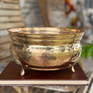 May include: A polished brass bowl with a rounded body and a wide, open top. The bowl features a smooth, reflective surface and is supported by three small, curved metal feet. It rests on a flat, rectangular, dark brown book cover.