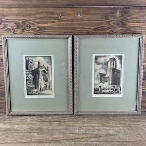 May include: Two framed prints featuring black and white architectural sketches. The left print depicts a gothic cathedral, while the right print shows a large skyscraper near a bridge. Both are encased in silver textured frames with sage mats.