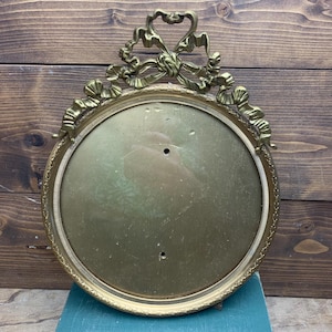 May include: A round brass picture frame featuring an ornate bow-shaped crest at the top. The frame has a textured, beaded inner border and a smooth, reflective gold-toned metal backing plate with two small circular holes positioned vertically.
