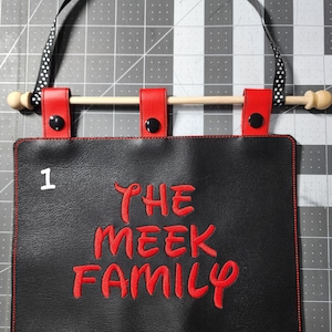 May include: Black leather banner with red stitching and a red embroidered phrase "THE MEEK FAMILY". The banner has a wooden dowel rod and black and white polka dot ribbon.