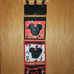 May include: A black fabric wall organizer with three pockets featuring Mickey Mouse designs. The top pocket is plain black, the middle pocket has a white background with Mickey Mouse heads in red and black, and the bottom pocket has a black background with Mickey Mouse heads in red and white. Each pocket has a red border and two metal rings.