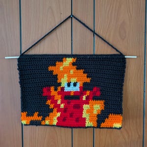 May include: A handmade, crocheted wall hanging featuring a pixelated design of a red and orange creature against a black background. The artwork is suspended by a black yarn and a wooden dowel.