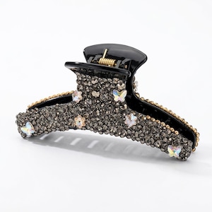 May include: A black hair claw clip featuring a dense surface of dark gray rhinestones. The clip is embellished with several small, iridescent butterfly-shaped accents and has a gold-toned metal spring mechanism at the top hinge.