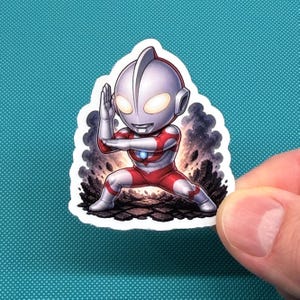 Op de afbeelding: Een sticker met een chibi-stijl karakter in een rood en wit pak, in een vechtkunsthouding. Het karakter heeft een zilveren helm en gloeiende gele ogen. De achtergrond toont een rokerige, rotsachtige sc&egrave;ne.