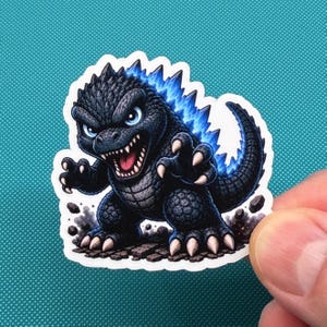 Puede incluir: Una pegatina con un Godzilla de dibujos animados. La criatura es principalmente negra con detalles azules en sus aletas dorsales y un contorno blanco. Su boca está abierta, revelando dientes.
