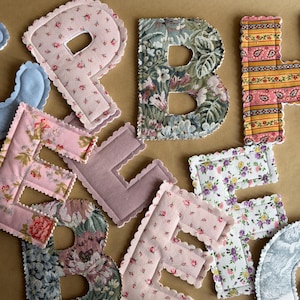 Peut inclure: Une collection de lettres en tissu présentant divers motifs, notamment des imprimés floraux roses, du mauve uni, des rayures cachemire jaunes et des designs en tapisserie complexes. Chaque lettre a un bord festonné et rembourré.