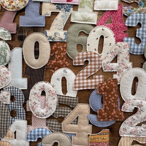 May include: A collection of fabric numbers featuring various patterns including floral, gingham, plaid, and solid colors. Each number is individually cut and stitched with a scalloped edge, displaying a diverse array of textile designs.