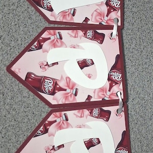 Peut inclure: Une banderole décorative composée de cinq fanions pentagonaux sur fond rose clair, ornés d'un motif de bouteilles de Dr Pepper et de nœuds roses. Chaque fanion porte une grande lettre blanche, formant ensemble le mot HAPPY, reliés entre eux.