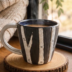 May include: A black and white ceramic mug with a birch tree design, filled with a warm beverage. The mug sits on a round wooden coaster. A cozy, cream-colored sweater is in the background, near a window.