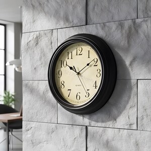 May include: A round wall clock with a black frame and a cream-coloured face. The clock features black Arabic numerals from 1 to 12, black hour, minute, and second hands, and the word QUARTZ printed in black text below the centre of the clock face.