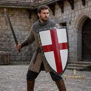 Puede incluir: Un escudo medieval tipo heater con una cruz roja sobre fondo blanco, bordeado por un marco metálico oscuro con remaches. Se presenta junto a una espada larga de acero, una cofia de cota de malla, un gambesón acolchado y botas de cuero.
