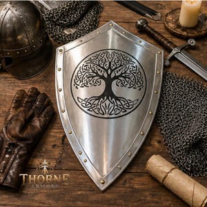 Puede incluir: Un escudo de metal plateado con un diseño circular negro del árbol de la vida con intrincados patrones de nudos. El escudo tiene una base puntiaguda y está decorado con pequeños remaches de color latón a lo largo del borde exterior pulido.