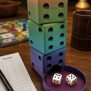 May include: A vertical dice tower constructed from three stacked cubes featuring a gradient color transition from peach to green to blue. The base includes a circular purple tray containing two standard white six-sided dice with black pips.