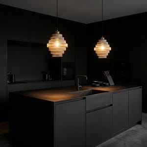 Könnte beinhalten: Zwei Pendelleuchten mit einem geschichteten, zylindrischen Design hängen über einer schwarzen Kücheninsel. Die Leuchten haben einen warmen, bernsteinfarbenen Schein. Die Küche verfügt über schwarze Schränke, ein Waschbecken und eine Armatur.