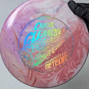May include: A pink and white disc golf disc with the text "CHRIS CLEMONS" and "JAWBREAKER ESP METEOR" in a rainbow holographic design. The disc has a marbled appearance.