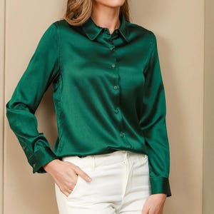 May include: A long-sleeved emerald green button-down shirt with a classic collar and a smooth, satin-like finish. The shirt features matching green buttons down the front placket and at the cuffs, paired with plain white trousers.