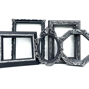 May include: A collection of four empty picture frames in various shapes including rectangular, oval, and octagonal. Each frame features a matte black finish with intricate silver-toned embossed scrollwork and floral detailing along the inner borders.