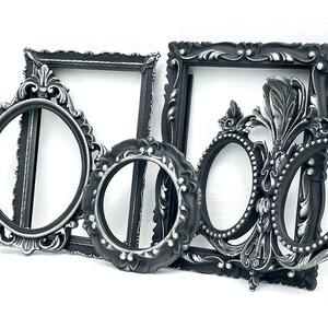 May include: A collection of four ornate picture frames in matte black with silver highlights. The set includes two rectangular frames and two oval frames, all featuring intricate raised scrollwork and decorative detailing along the edges.