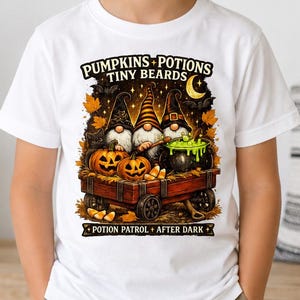 May include: A white short-sleeve t-shirt featuring a graphic print of three gnomes in a wooden wagon with pumpkins and a bubbling green cauldron. The text reads PUMPKINS POTIONS TINY BEARDS and POTION PATROL AFTER DARK in black lettering.