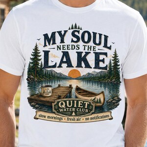 May include: A white short-sleeve t-shirt featuring a graphic of a lake, a wooden dock, a canoe, and a coffee mug. The text reads: MY SOUL NEEDS THE LAKE, QUIET WATER CLUB, slow mornings, fresh air, no notifications, and LIFE IS BETTER HERE.