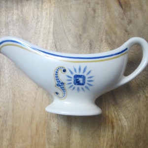 May include: White ceramic gravy boat with a handle and spout. It features blue and gold trim along the rim and a decorative blue and gold design on the side. The gravy boat sits on a wooden surface.