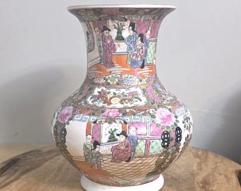 Op de afbeelding: Een keramische vaas met een uitlopende rand en een bolvormig lichaam, versierd met handgeschilderde scènes van figuren in traditionele kleding, bloemmotieven en geometrische patronen in roze, groen, oranje en zwart op een witte achtergrond.
