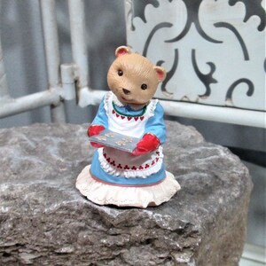 May include: A figurine of a bear wearing a blue dress and a white apron with red accents, holding a tray of cookies. The bear has a light brown face and is standing on a rock.