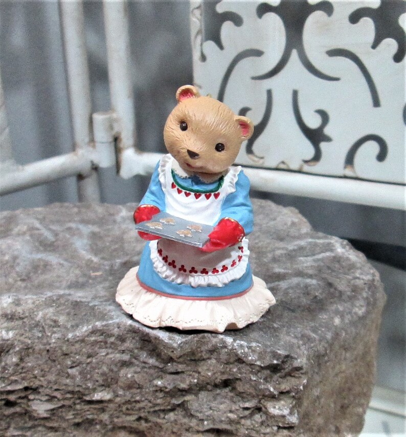 May include: A figurine of a bear wearing a blue dress and a white apron with red accents, holding a tray of cookies. The bear has a light brown face and is standing on a rock.