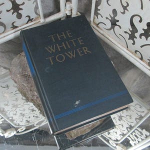 May include: A dark blue hardback book with the title "THE WHITE TOWER" in gold lettering. The book is resting on a white metal surface with a decorative design.