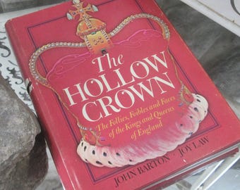 Op de afbeelding: Een rood hardcover boek met de titel "The Hollow Crown" met een kroonillustratie. De titel staat in grote witte letters, met de namen van de auteurs onderaan. Het boek gaat over de koningen en koninginnen van Engeland.