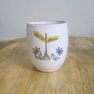 May include: White ceramic tumbler with a hand-painted design. The design includes a yellow signpost pointing to Luzern and Bern, with colorful flowers at the base. The tumbler has a slightly flared shape.