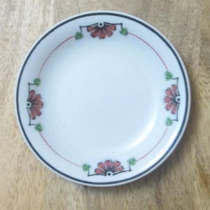 May include: A small, white ceramic plate with a black rim and a red and green floral design. The plate has a simple, elegant design, perfect for serving appetizers or desserts. The plate is approximately 6 inches in diameter.