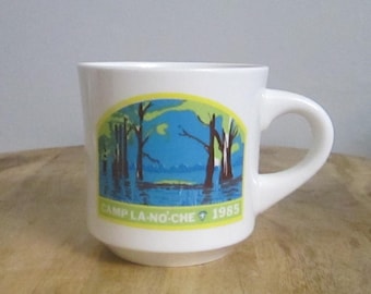 Könnte beinhalten: Weiße Keramik-Tasse mit gebogenem Henkel. Die Tasse zeigt eine farbenfrohe Grafik einer Landschaft mit Bäumen und Wasser sowie den Text "CAMP LA-NO-CHE + 1985". Die Tasse steht auf einer Holzoberfläche.