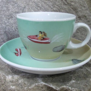 May include: A green and white ceramic teacup and saucer set with a cartoon illustration of two people riding a red jet ski on a turquoise body of water. The saucer has a red and white shoe design on it.